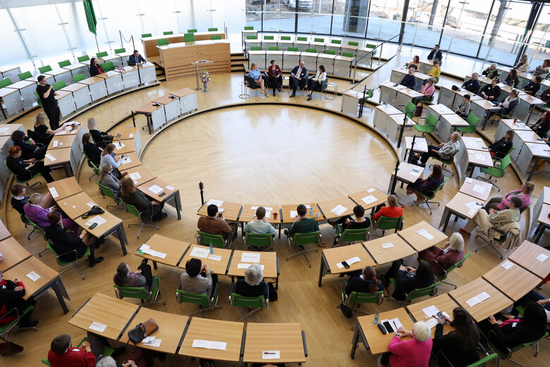 Weitwinkelaufnahme des Plenarsaals des Sächsischen Landtags. Im Zentrum des Saales sitzen die Sozialministerin und drei Gesprächspartner.